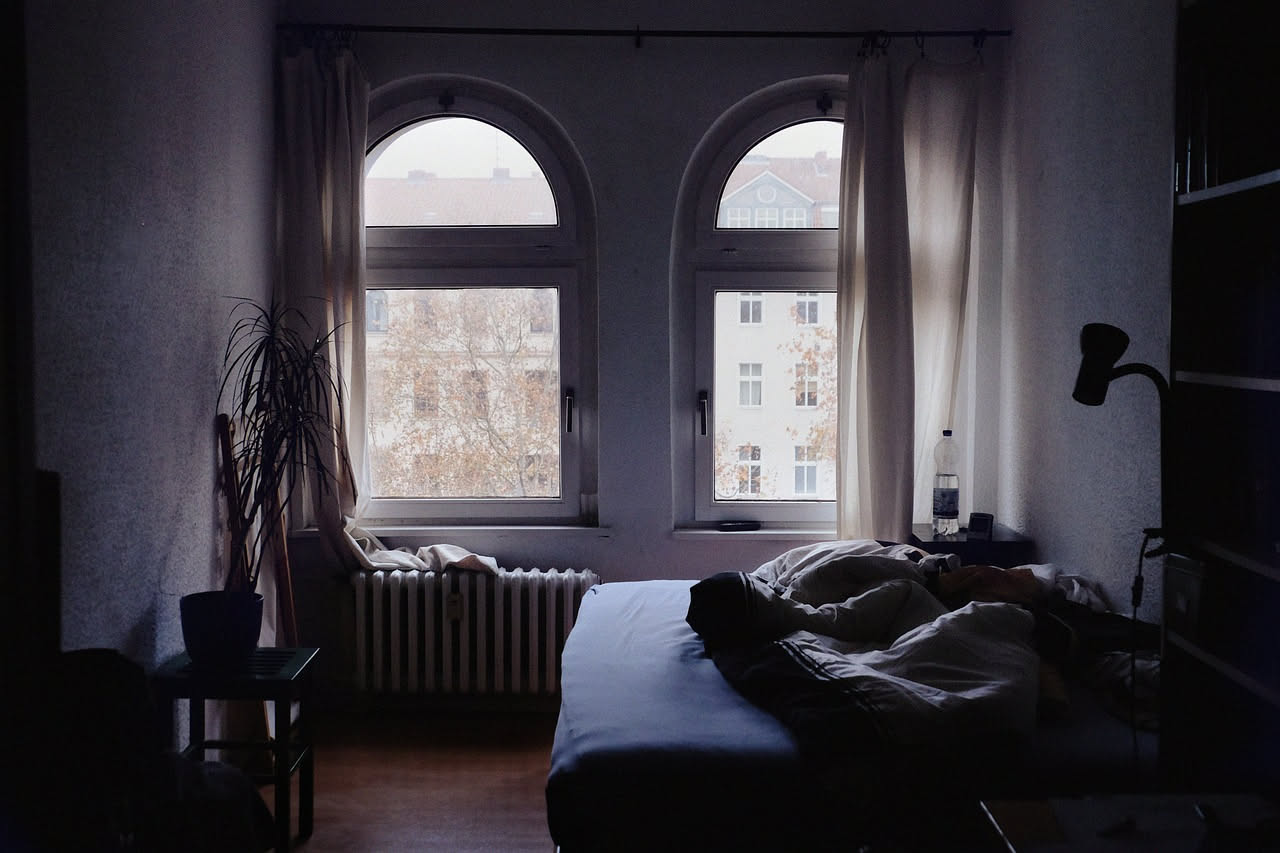 Camera da letto con finestra aperta al tramonto, tende bianche che ondeggiano per la brezza, letto con coperta chiara e cuscini bianchi