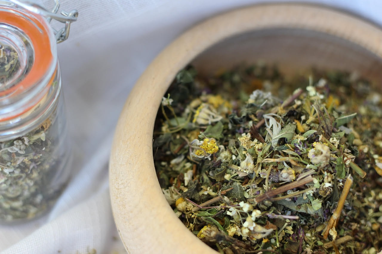 Tazza di ceramica bianca con tisana calda fumante, fiori di camomilla secchi, bustine di tè alle erbe e un libro aperto su un tavolo di legno scuro in penombra
