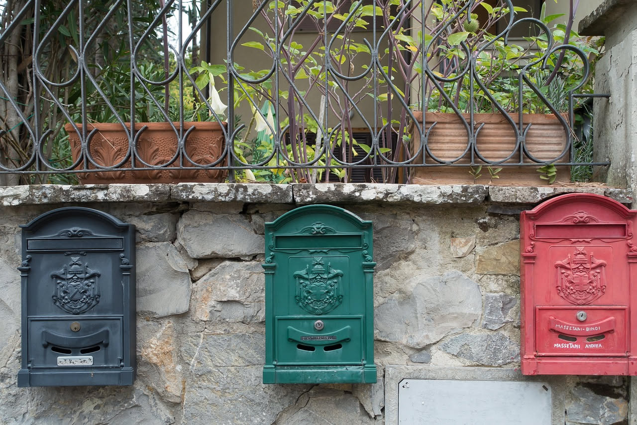Una cassetta postale rossa tipica italiana fissata a un muro, leggermente corrosa dal tempo, con la bocca di inserimento in primo piano