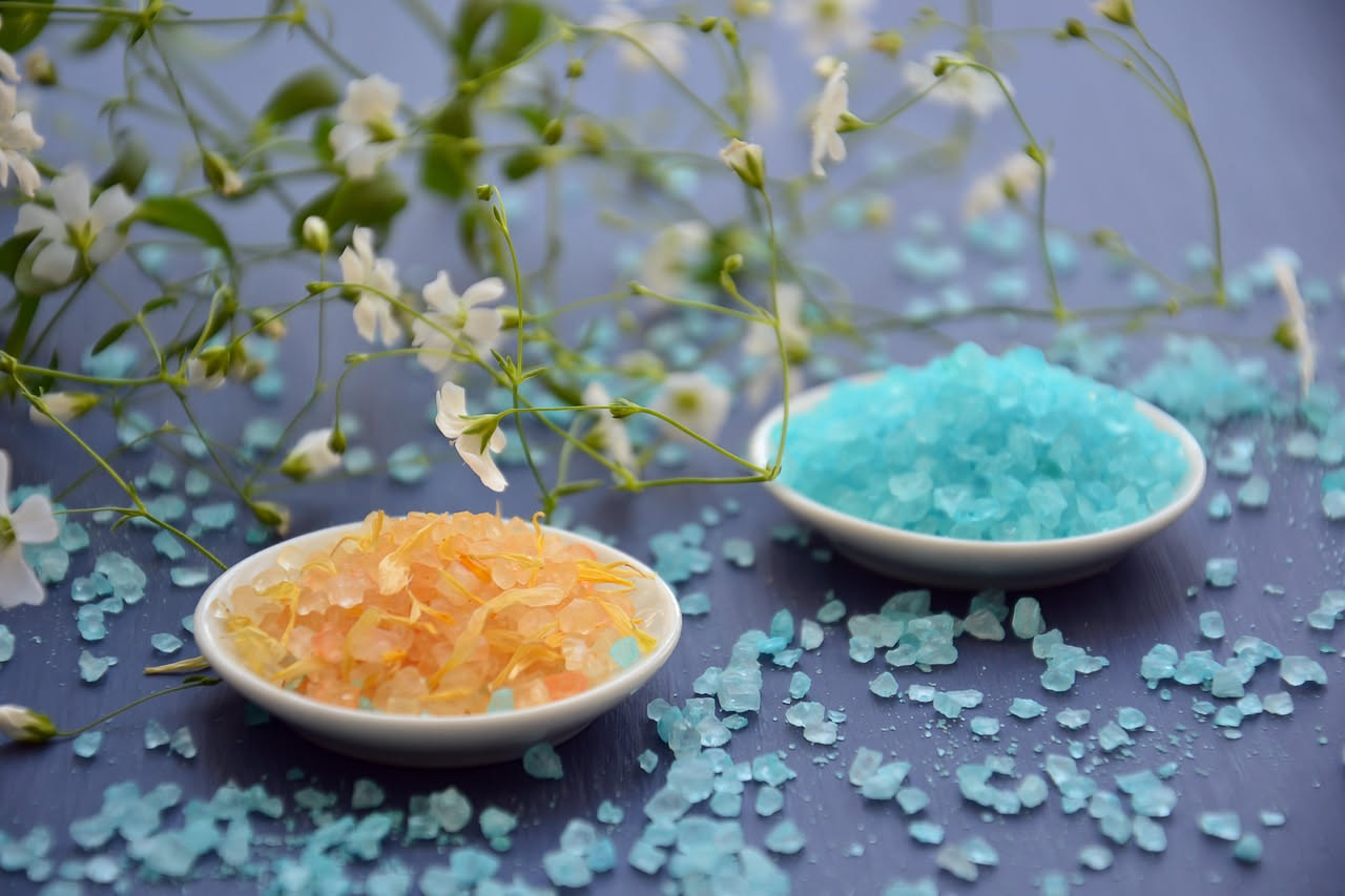 Vasca da bagno riempita di acqua calda con cristalli di sale grosso sparsi sul bordo bianco, accanto asciugamani grigio chiaro e una bottiglia di olio essenziale