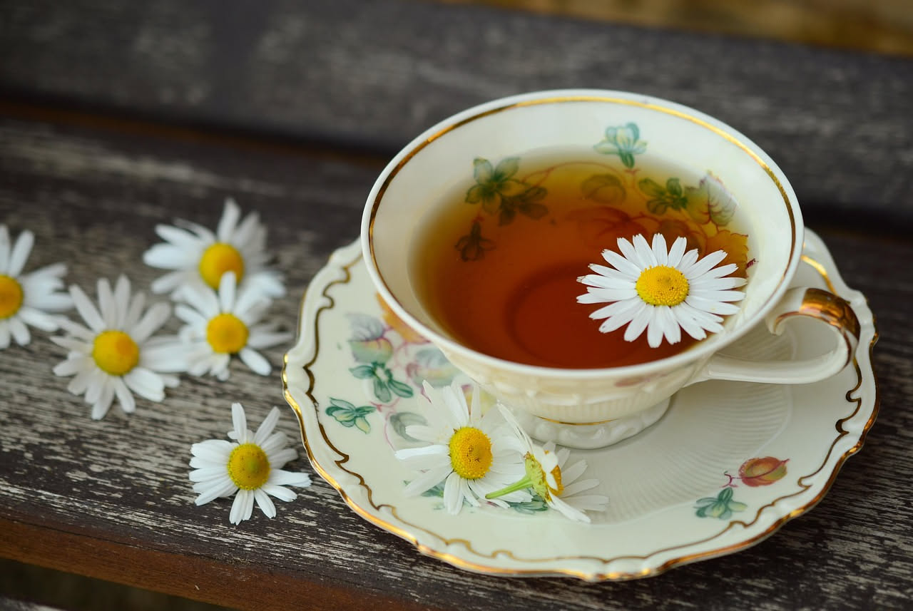 Tazza di ceramica bianca con camomilla in infusione, fiori di camomilla secchi sparsi attorno, vapore caldo che sale, luce naturale mattutina su sfondo sfocato