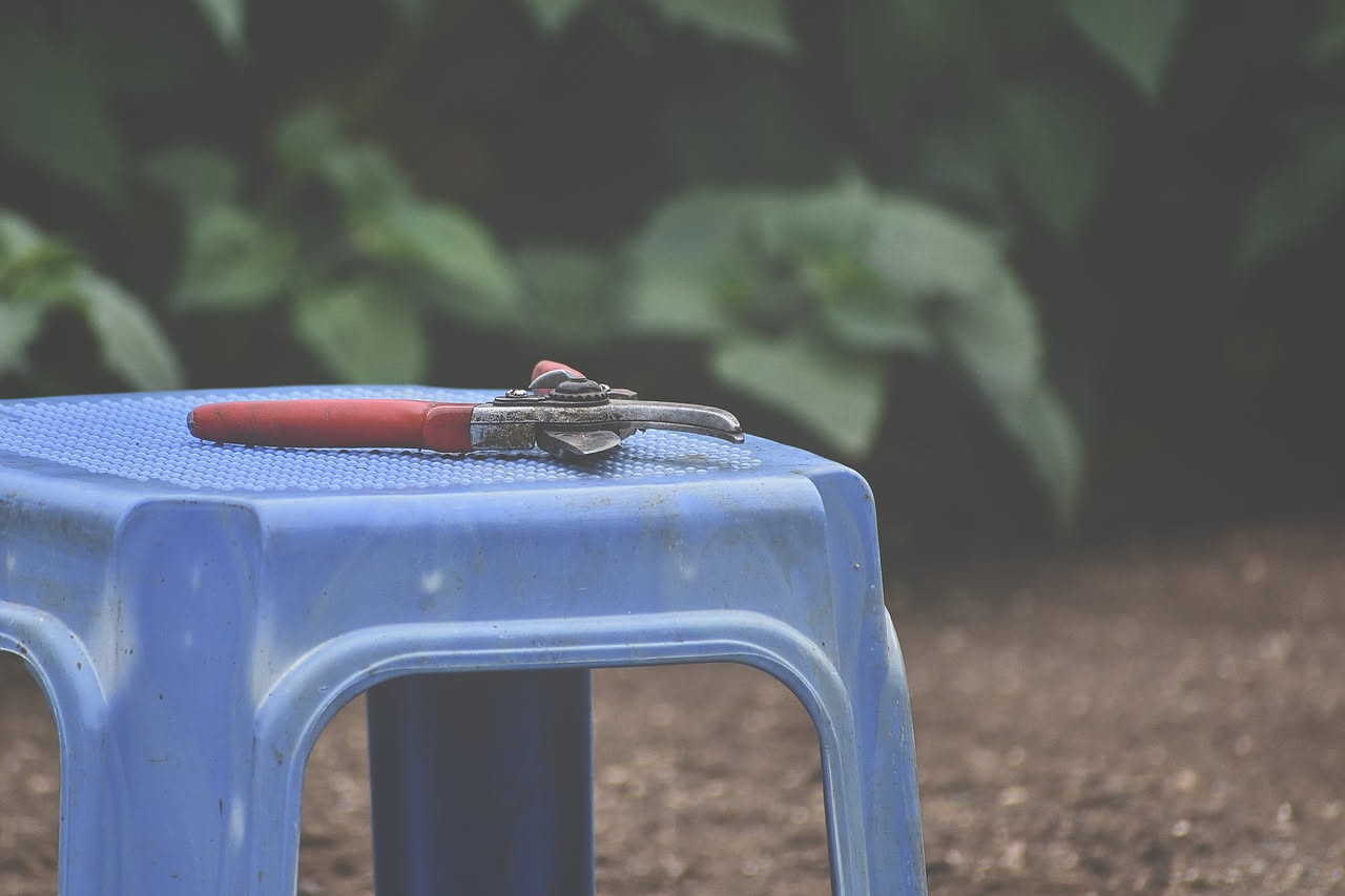 Sedia da giardino in plastica grigia con crepa sulla spalliera, vicino un phon blu e un rotolo di nylon trasparente su un tavolo di legno