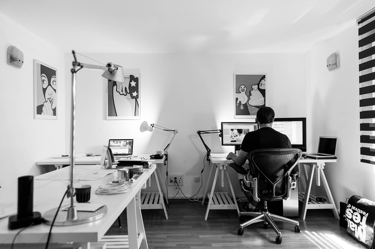Una stanza moderna con scrivania bianca, sedia da ufficio grigia, finestra con luce naturale e parete decorata, allestita come uno spazio di lavoro condiviso in ambiente residenziale