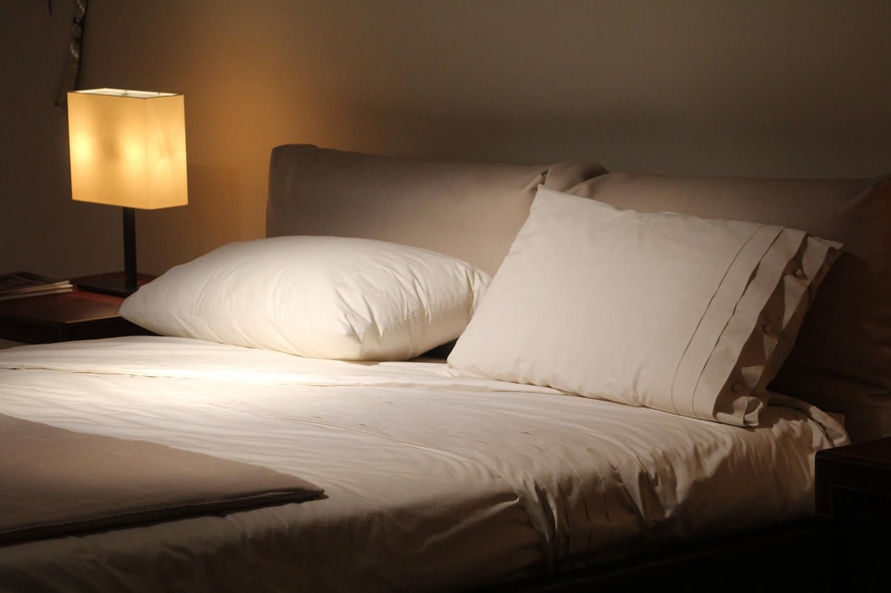 Camera da letto minimalista con letto matrimoniale in legno chiaro, lenzuola bianche in cotone, cuscini morbidi, persiane chiuse, lampada dimmerabile e umidificatore bianco su comodino