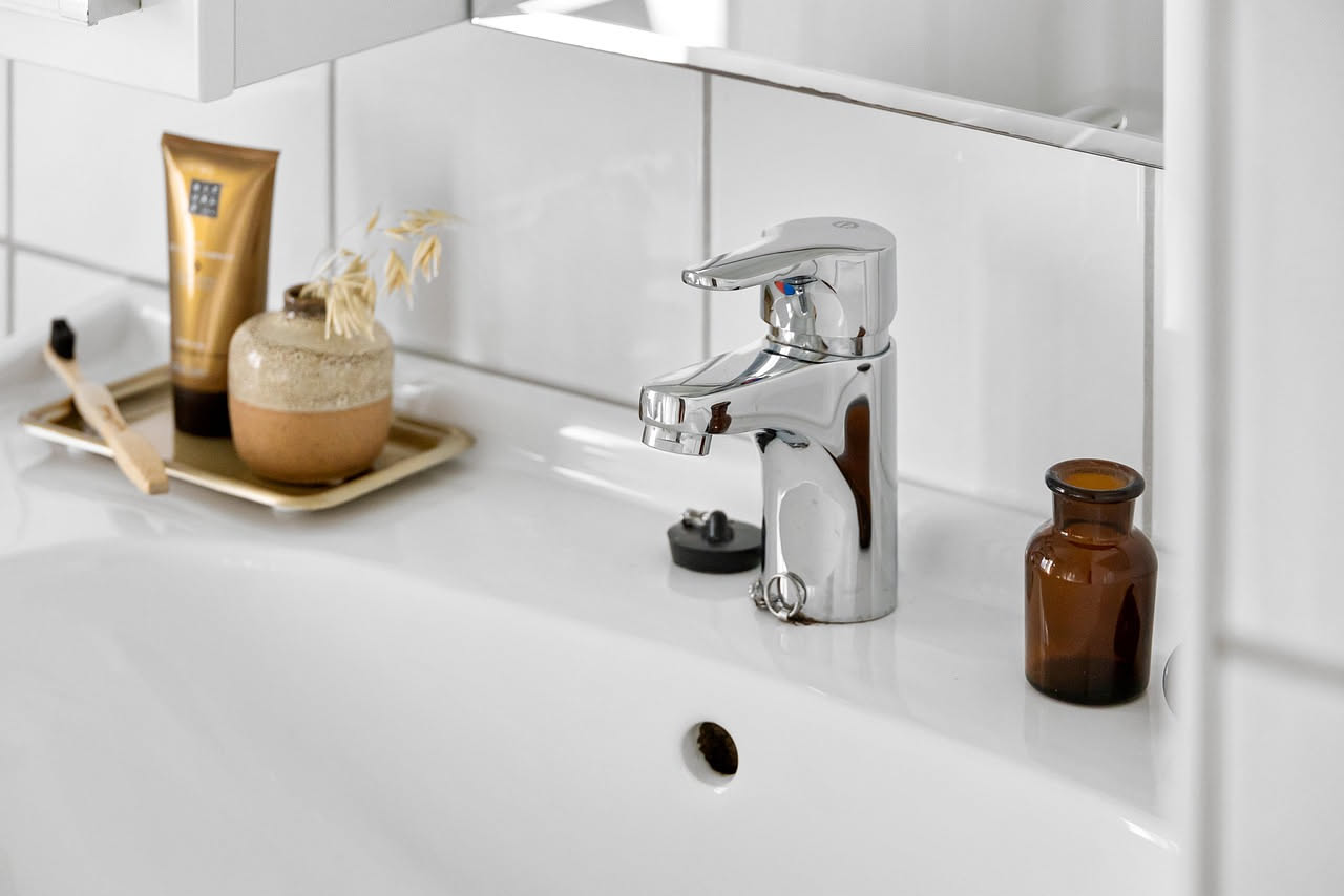 Mano con cacciavite che smonta il tappo di un lavabo bianco, con chiave inglese e nuovo tappo di ricambio appoggiati sul ripiano